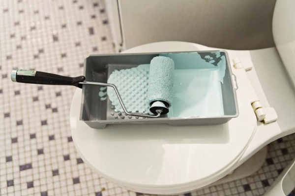 Roller and tray filled with light blue paint resting on top of toilet bowl.